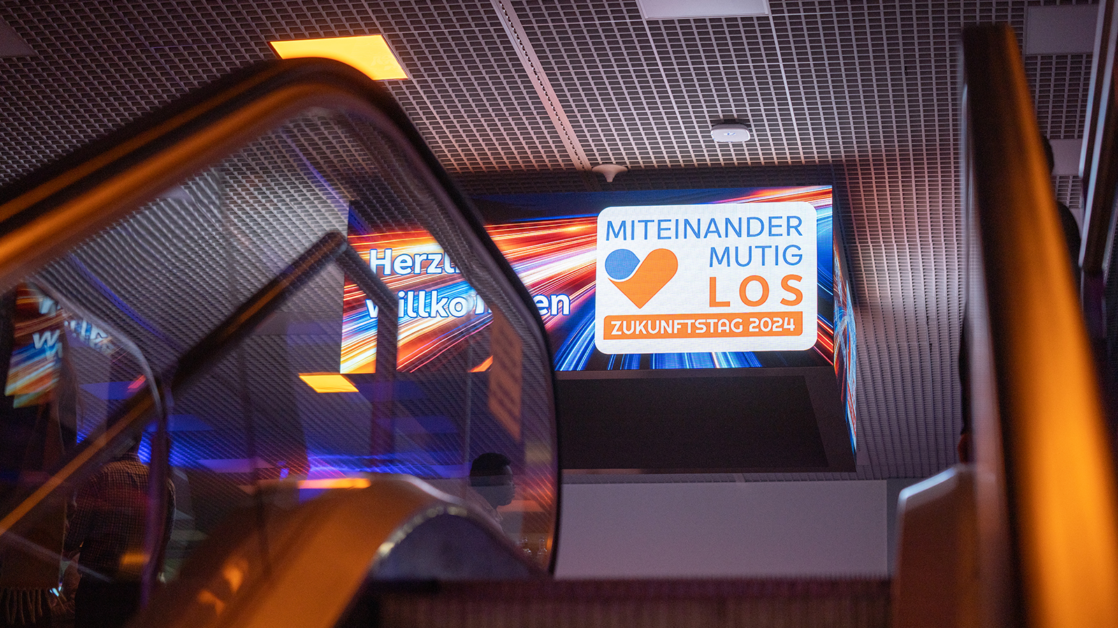 Leuchttafel mit dem Motto „Miteinander. Mutig. Los. Zukunftstag“ über einer Rolltreppe